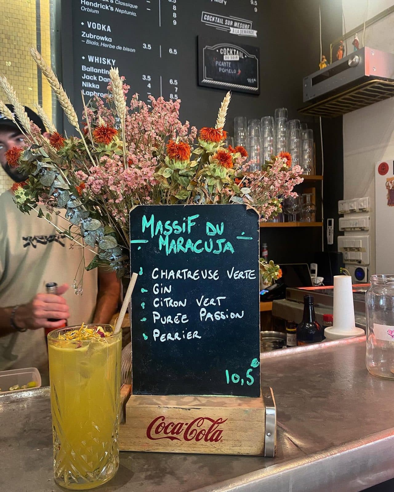 Un cocktail et son ardoise manuscrite devant les fleurs séchées du comptoir.