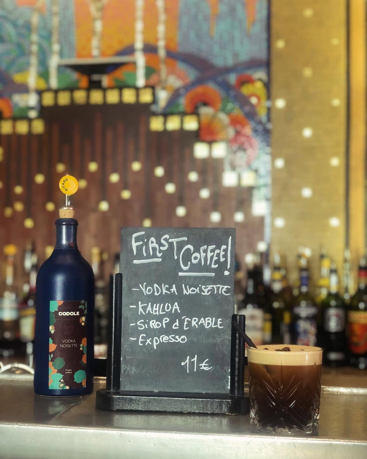 Un café-cocktail posé sur le zinc, l’ardoise du jour et la mosaïque en arrière-plan.
